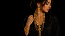 A woman wearing an ornate gold-toned layered necklace, matching drop earrings, and stacked bangles against a solid black background.