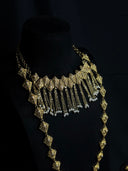 Close-up of a gold-tone diamond tile choker necklace on a black bust, showing engraved tile links and cascading chain tassels tipped with pearl-like beads