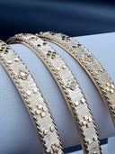 Close-up of gold-tone Barocco-style bangles showing rigato line-engraved texture, raised polished floral motifs, and milgrain beaded border