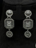 Close-up of the Turath three-tier drop earrings showing the round scrollwork stud top, square filigree medallion with engraved floral centre, and round disc drop